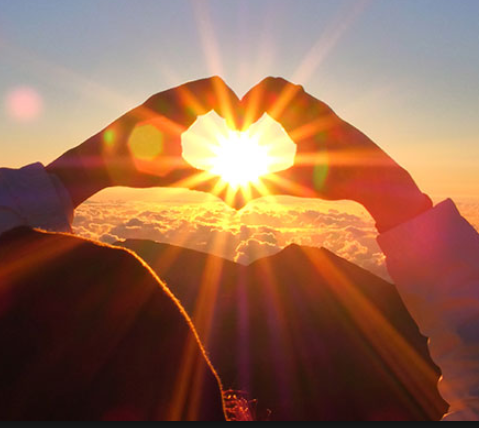 person standing before mountains at sunset just as sun approaches horizon line, she is holding her hands up in the shape of a heart so that the sun rays pass through the heart made of her hands